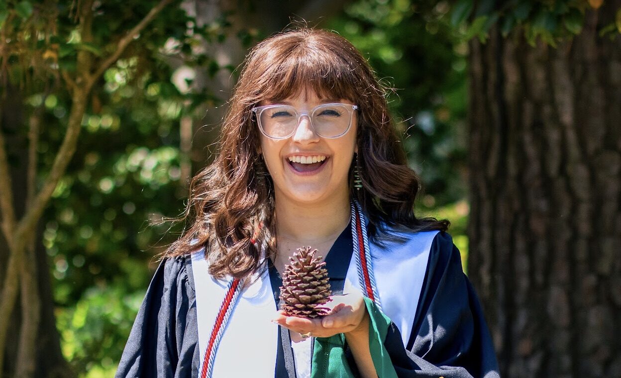 Emma wearing a graduation gown and holding a pine cone.